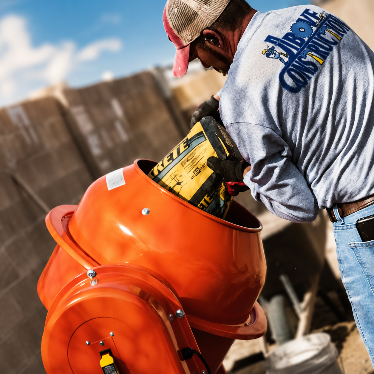 Above Construction worker pouring cement mix into an orange mixer on a sunny job site